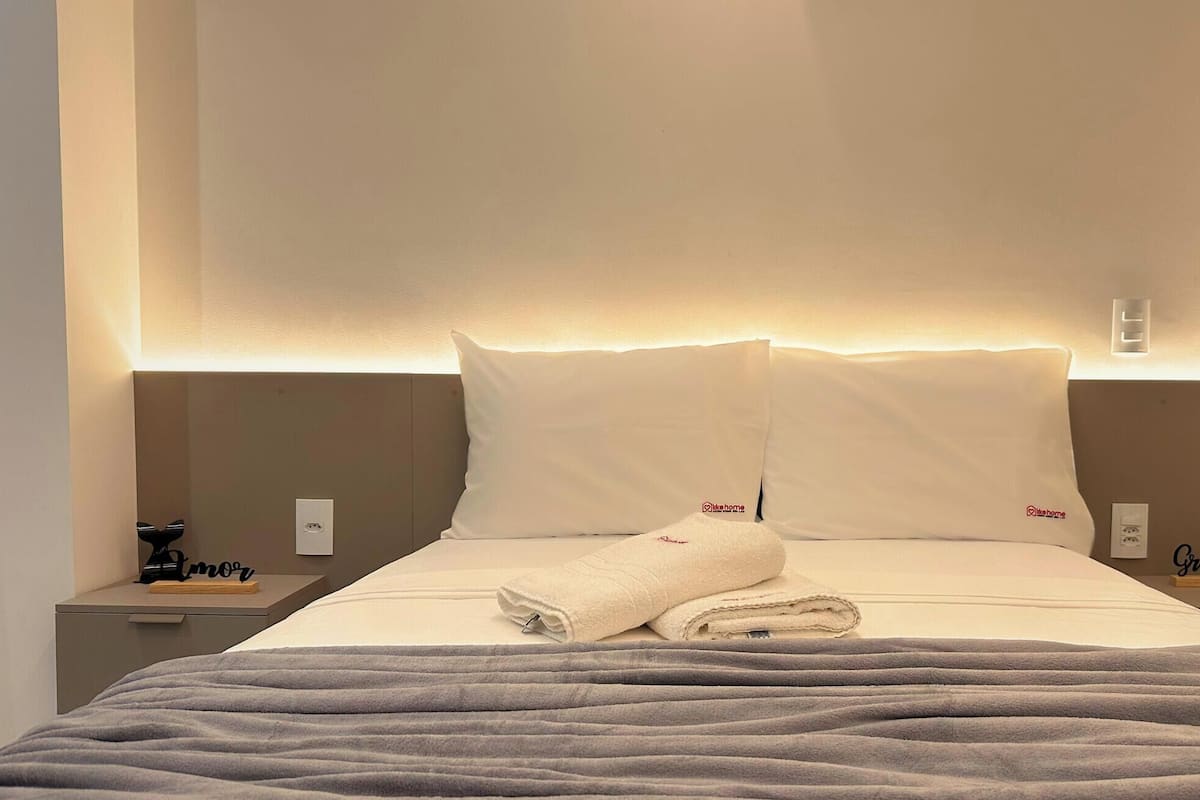 Uma cama bem arrumada com uma toalha dobrada, uma mesa de cabeceira com um telefone e um objeto pequeno, e uma luminária fixada na parede.