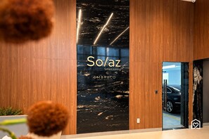 A modern entrance with a wooden panel wall, a black marble wall with text, and a glass door leading to a parking area. - Solaz - LikeHome Hospedagens (João Pessoa)