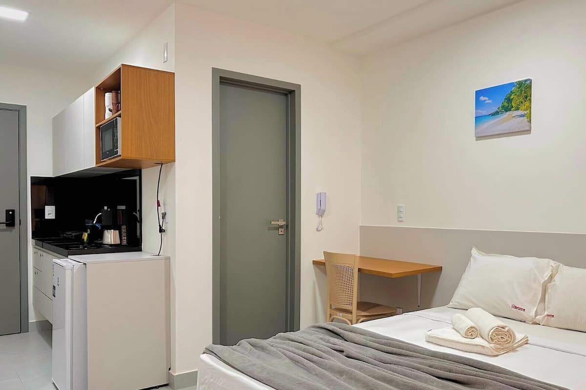 Quarto de hotel com cama, escrivaninha, cozinha compacta e porta de acesso a outro cômodo.