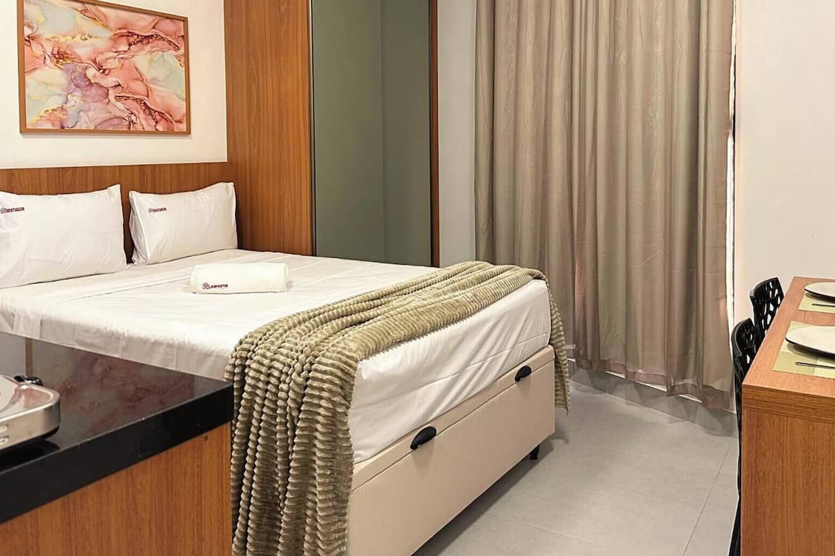 Quarto de hotel com cama, escrivaninha com laptop, cafeteira e um quadro na parede.