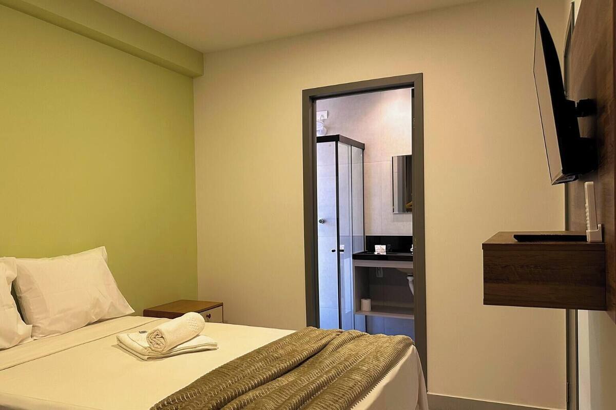 Quarto de hotel com uma cama, uma televisão na parede e uma porta para o banheiro.