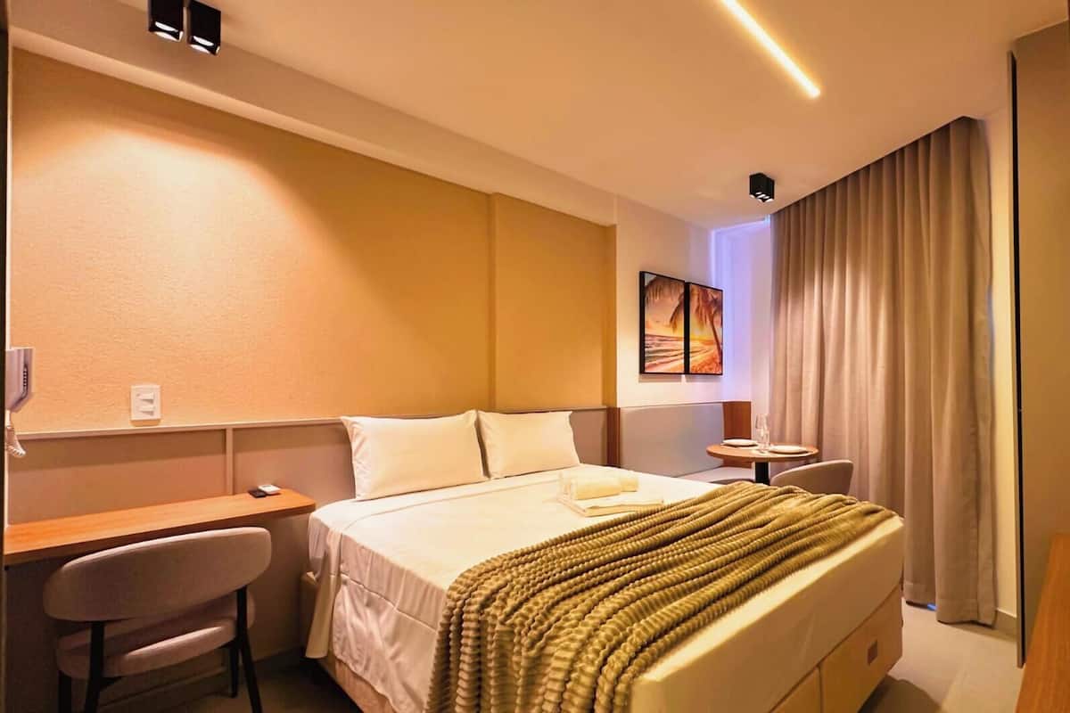Quarto de hotel com cama, escrivaninha, cadeira, mesa pequena com um copo e um quadro na parede.