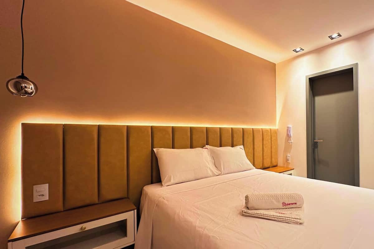 Quarto de hotel com uma cama grande, mesa de cabeceira, luminária na parede e uma porta.