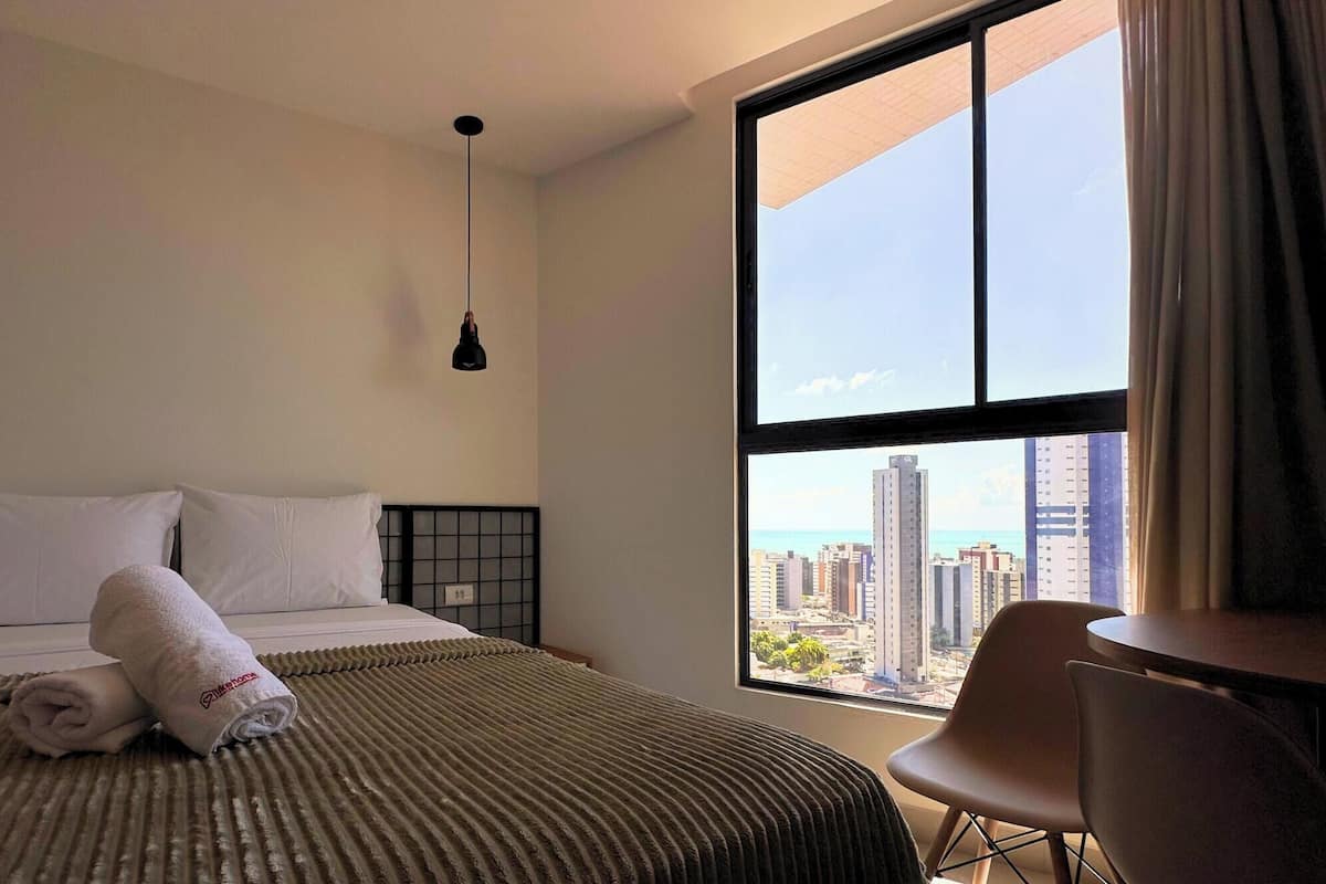 Quarto de hotel com uma janela ampla, uma cama com roupa de cama branca, uma toalha enrolada, uma mesinha e uma cadeira.