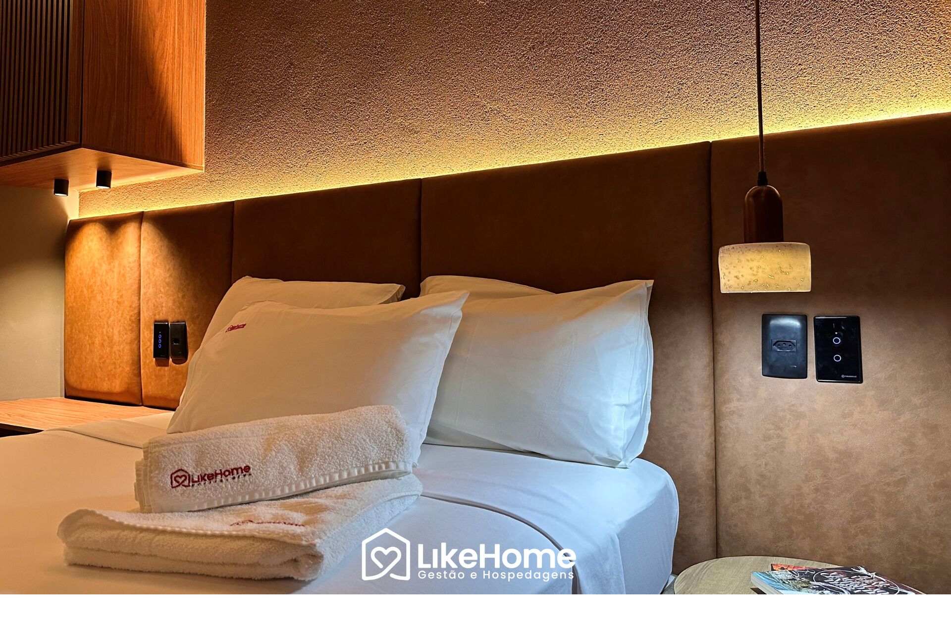 A hotel room with a bed, white bedding, a folded towel, a bedside table with a book, and a wall-mounted control panel.