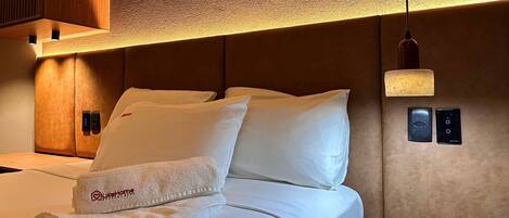 A hotel room with a bed, white bedding, a folded towel, a bedside table with a book, and a wall-mounted control panel.