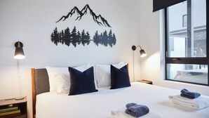 A neatly made bed with white and navy pillows, a mountain and forest wall art, and two bedside lamps. - Ripper Retreat - Modern Condo w/hot tub 5 minutes to Revelstoke Mountain Resort! (Revelstoke)