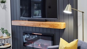 A modern living room with a flat-screen TV mounted on a dark wooden wall, a fireplace below, a blue sofa with a yellow pillow, a floor lamp, and a small round table with a plant. - Ripper Retreat - Modern Condo w/hot tub 5 minutes to Revelstoke Mountain Resort! (Revelstoke)