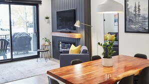 A modern dining area with a wooden table and chairs, a TV mounted on a wall, and a sliding glass door leading to an outdoor patio. - Ripper Retreat - Modern Condo w/hot tub 5 minutes to Revelstoke Mountain Resort! (Revelstoke)