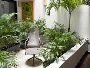 Un petit patio clos, avec une table ronde et une chaise en métal ajouré, entouré de différents pots de plantes et orné d’un grand tableau sur le thème de l’avocat.
