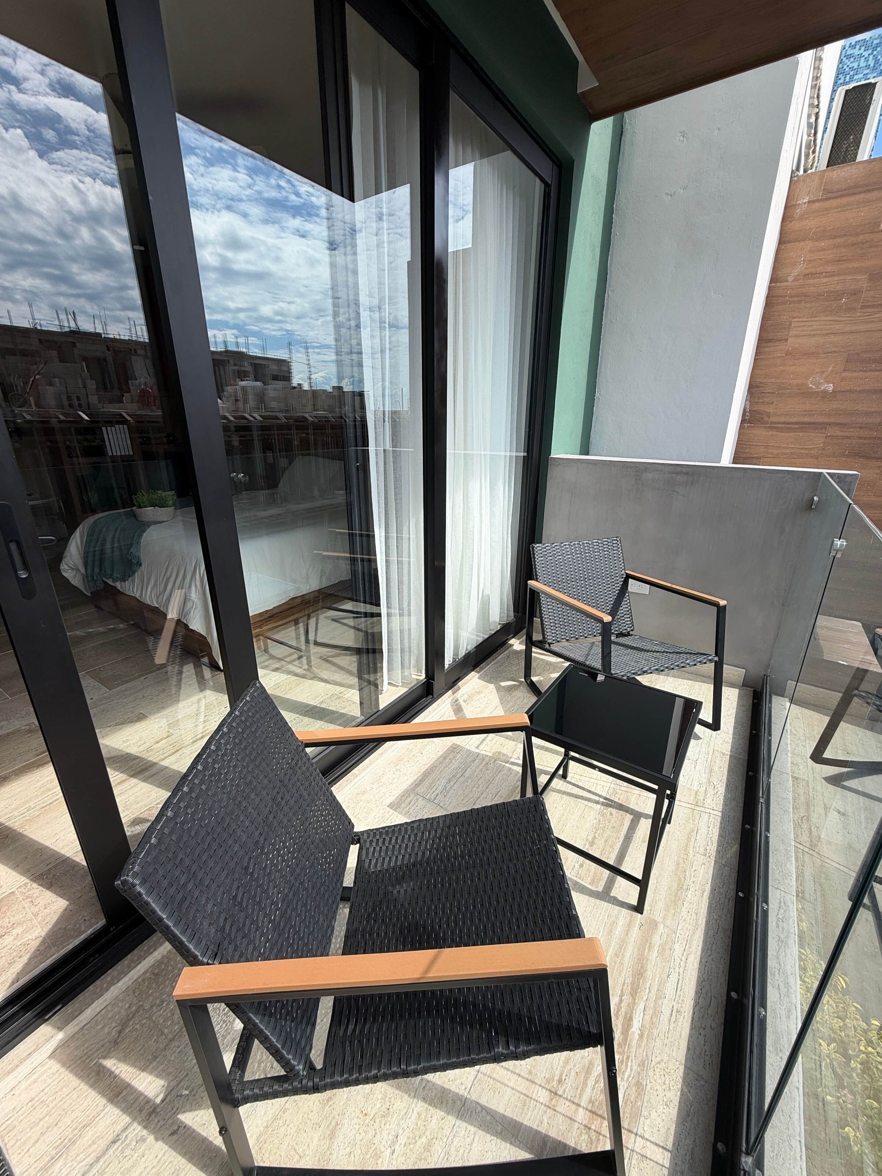A balcony with two wicker chairs and a glass table, large windows with sheer curtains, and a view of buildings in the background.