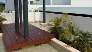 A modern outdoor pool area with wooden decking, a pergola, and various tropical plants. - Centro Playa Del Carmen Duna  508 (Playa del Carmen)