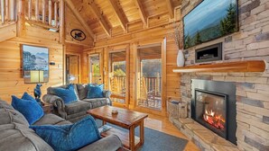 Ein Blockhaus-Interieur mit Kamin, Holzmöbeln und einem großen Fenster mit Blick auf eine Terrasse.