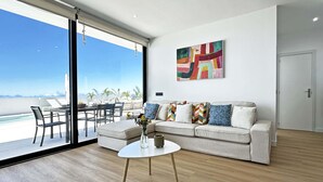 Een moderne woonkamer met een groot raam, een bank, een bijzettafeltje en een kleurrijk abstract schilderij.