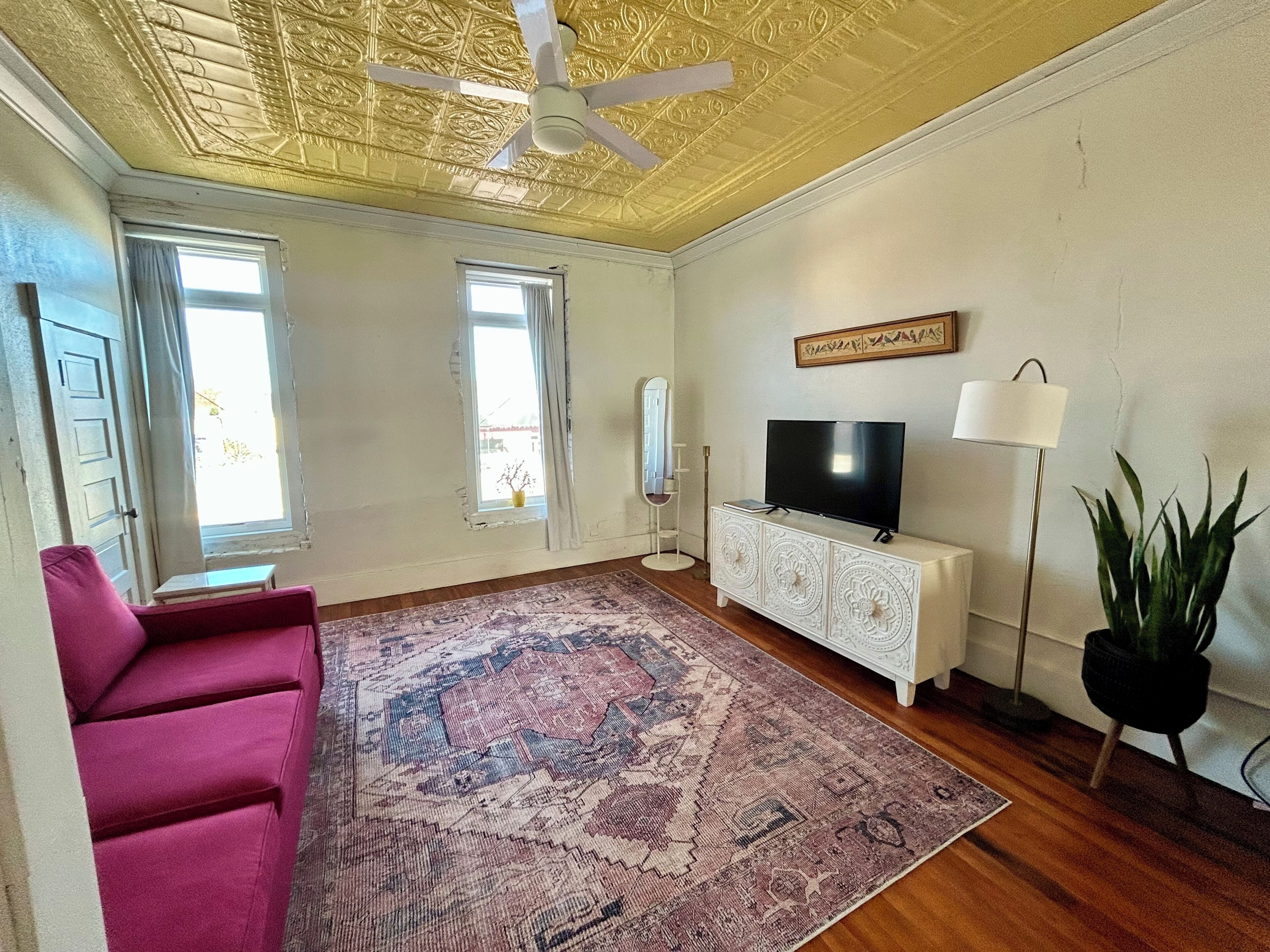 Ein Wohnzimmer mit Deckenventilator, einem Fernseher auf einem weißen Schrank, einem gemusterten Teppich, einem rosa Sofa und einer Topfpflanze.