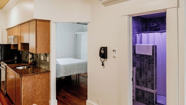 Een compacte keuken met houten kastjes, een magnetron en een gootsteen. In de aangrenzende kamer staat een klein bed en een badkamer met een douche en een handdoekrek.