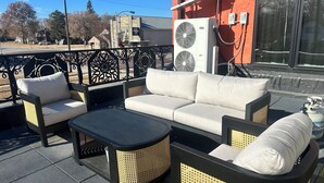 Een terras met een bank, stoelen en een tafel, naast een gebouw met airconditioningsapparatuur.