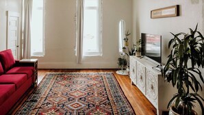 Een woonkamer met een rode bank, een kamerbreed tapijt met dessin, een wit tv-meubel en een grote plant.