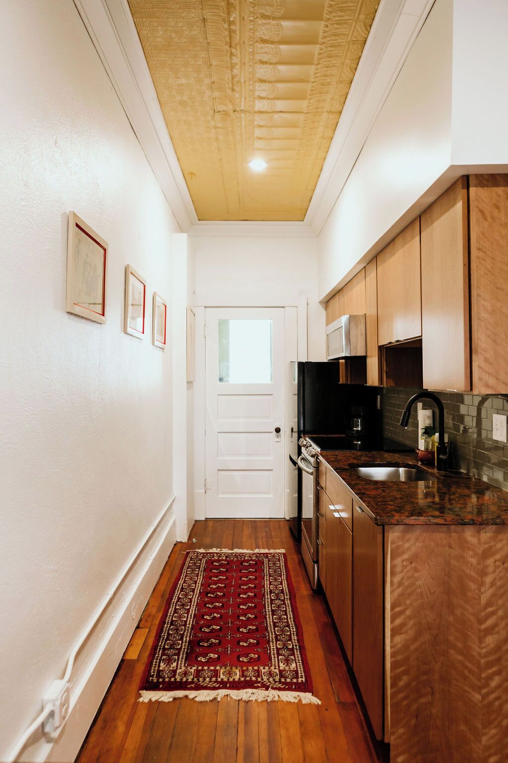 A narrow hallway leading to a kitchen with wooden cabinets, a microwave, and a stainless steel sink.