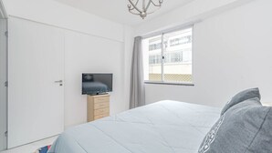Een slaapkamer met een bed, een televisie op een standaard, een raam met gordijnen en een kroonluchter.