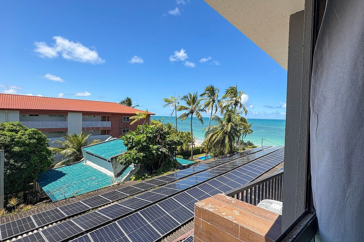A view of a hotel balcony overlooking a pool area with solar panels, palm trees, and a clear blue sky.