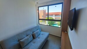 Une pièce avec un canapé, un téléviseur fixé au mur et une fenêtre donnant sur des immeubles et un arbre.