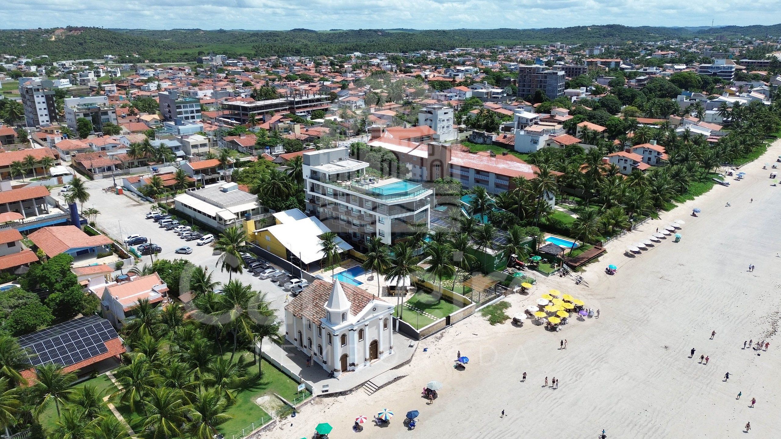 A coastal cityscape with a prominent hotel featuring a swimming pool, a church, and a beach with umbrellas.