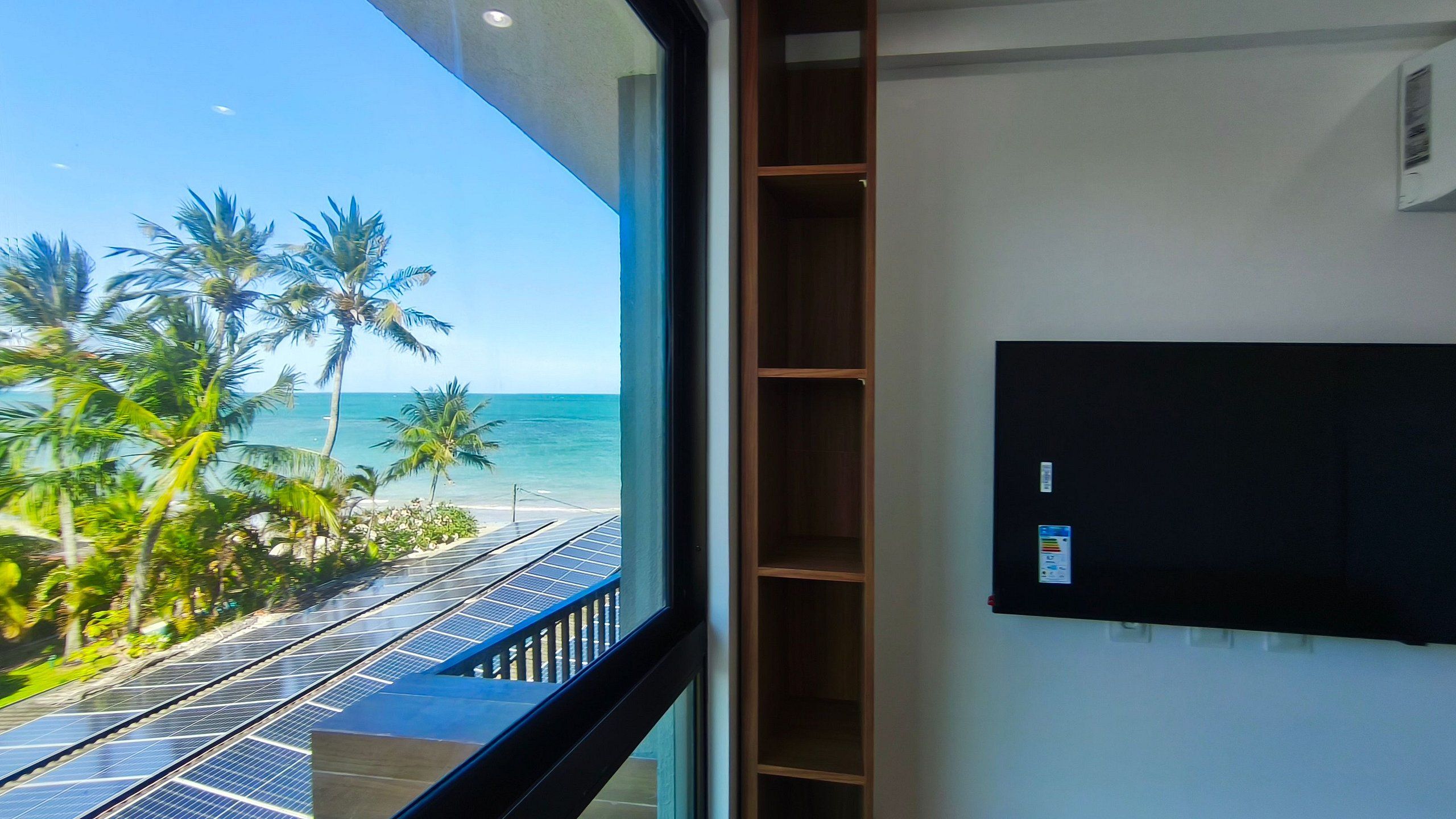 Una habitación con televisión montada en la pared, un balcón con paneles solares y vista al océano y palmeras.