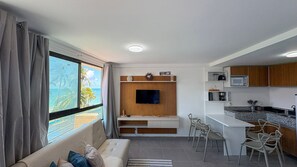 A compact living space with a sofa, a TV mounted on a wooden panel, a kitchenette with a microwave, and a dining area with chairs. - Portal dos Mares - Beira-Mar Tamandaré (Tamandaré)