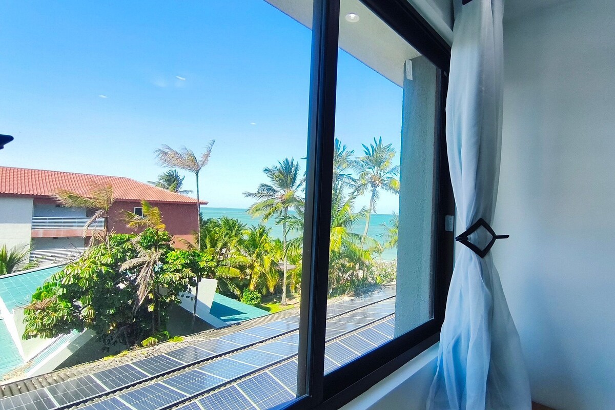 Una habitación con un amplio ventanal que da a una zona de piscina con paneles solares, palmeras y un edificio con techo rojo.