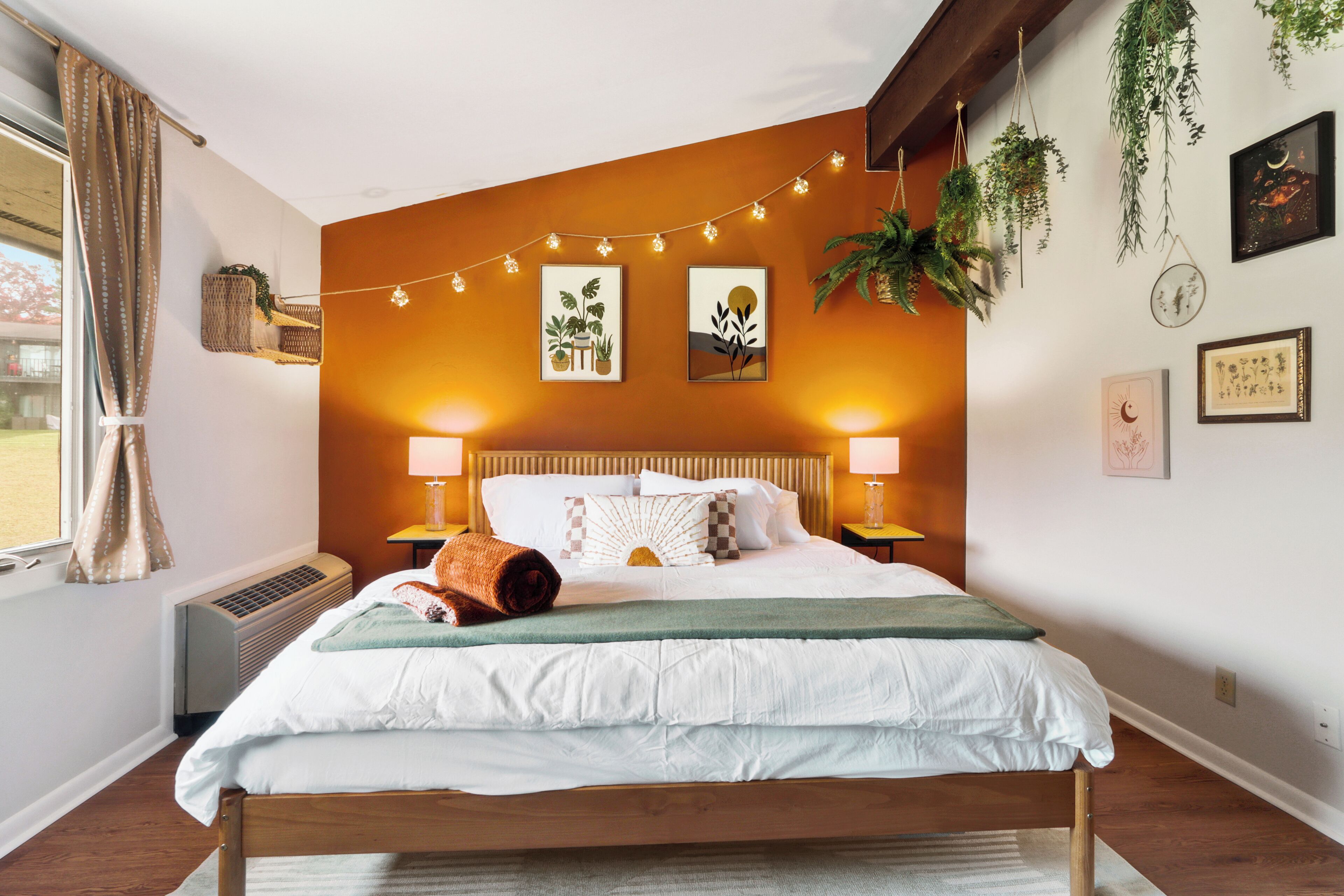 Ein Schlafzimmer mit einem Bett, Nachttischlampen, einem Fenster mit Vorhängen sowie Wanddekorationen, darunter gerahmte Bilder und Lichterkette.