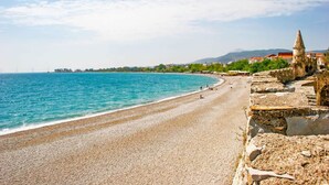 A coastal beach with a stone wall and a tower on the right. - Villa Psani Beachfront Escape With Garden (Nafpaktos)