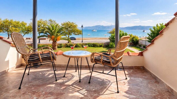 A balcony with two wicker chairs and a small round table, overlooking a coastal view with boats and greenery. - Villa Psani Beachfront Escape With Garden (Nafpaktos)