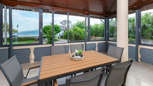 An outdoor dining area with a wooden table and chairs, large windows with a view of greenery and a body of water. - Villa Psani Beachfront Escape With Garden (Nafpaktos)