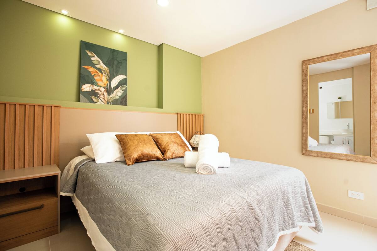 A neatly made bed with pillows, a bedside table, a wall-mounted mirror, and a painting.