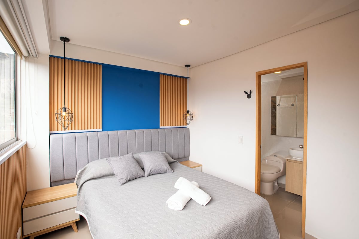 A modern bedroom with a bed, bedside tables, a headboard, and a bathroom with a toilet and sink.