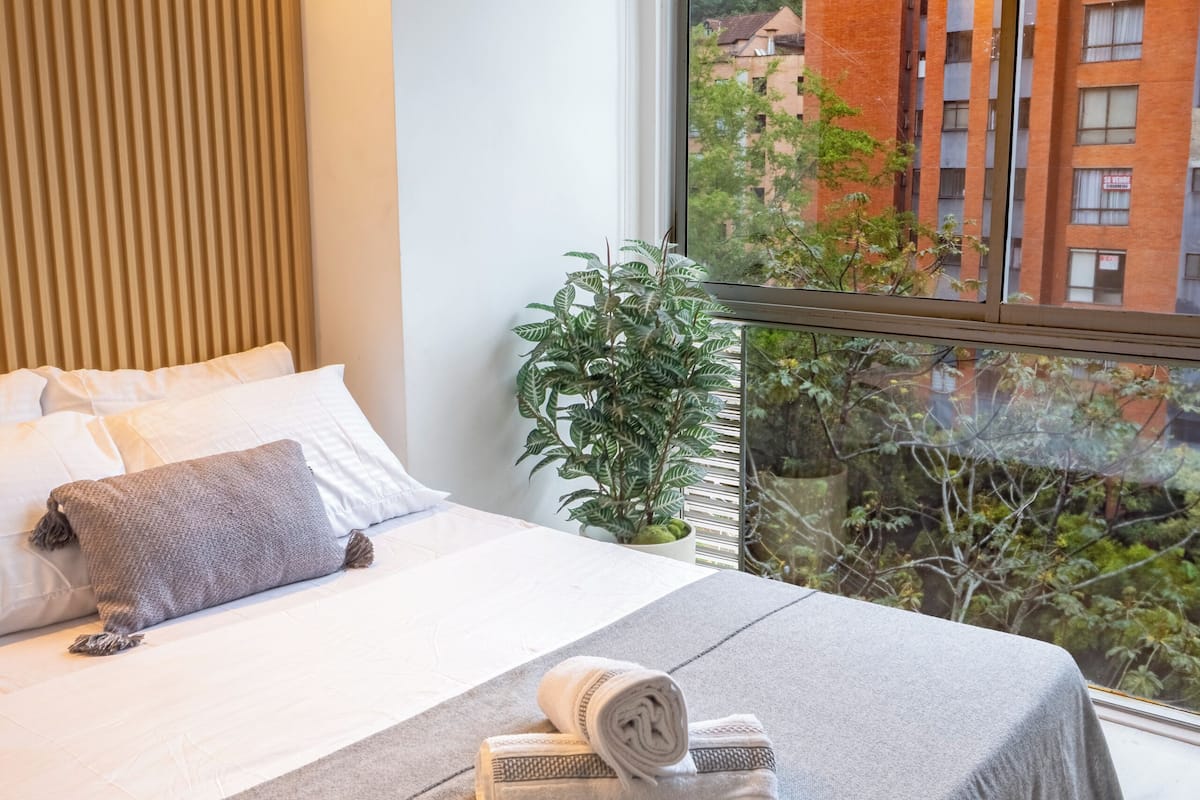 A modern hotel room with a large bed, a view of the city, and a balcony with a plant.