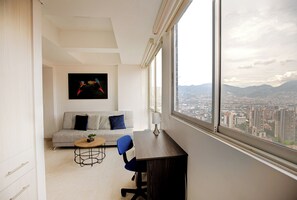 Un salon moderne avec un canapé, une table basse et un bureau offrant une vue sur le paysage urbain grâce à de grandes fenêtres.