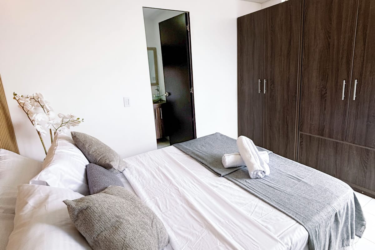 A bedroom with a bed, pillows, a bedside table, a wardrobe, and a bathroom visible through an open door.