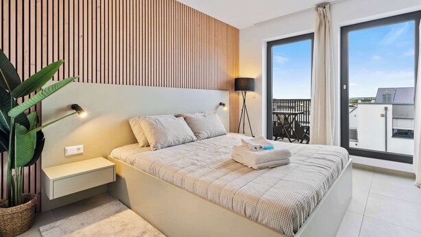 A modern bedroom with a large bed, bedside tables, a potted plant, and a balcony with a view. - Stylish Coliving Rooms for Work, Rest, and Play (Luxembourg)