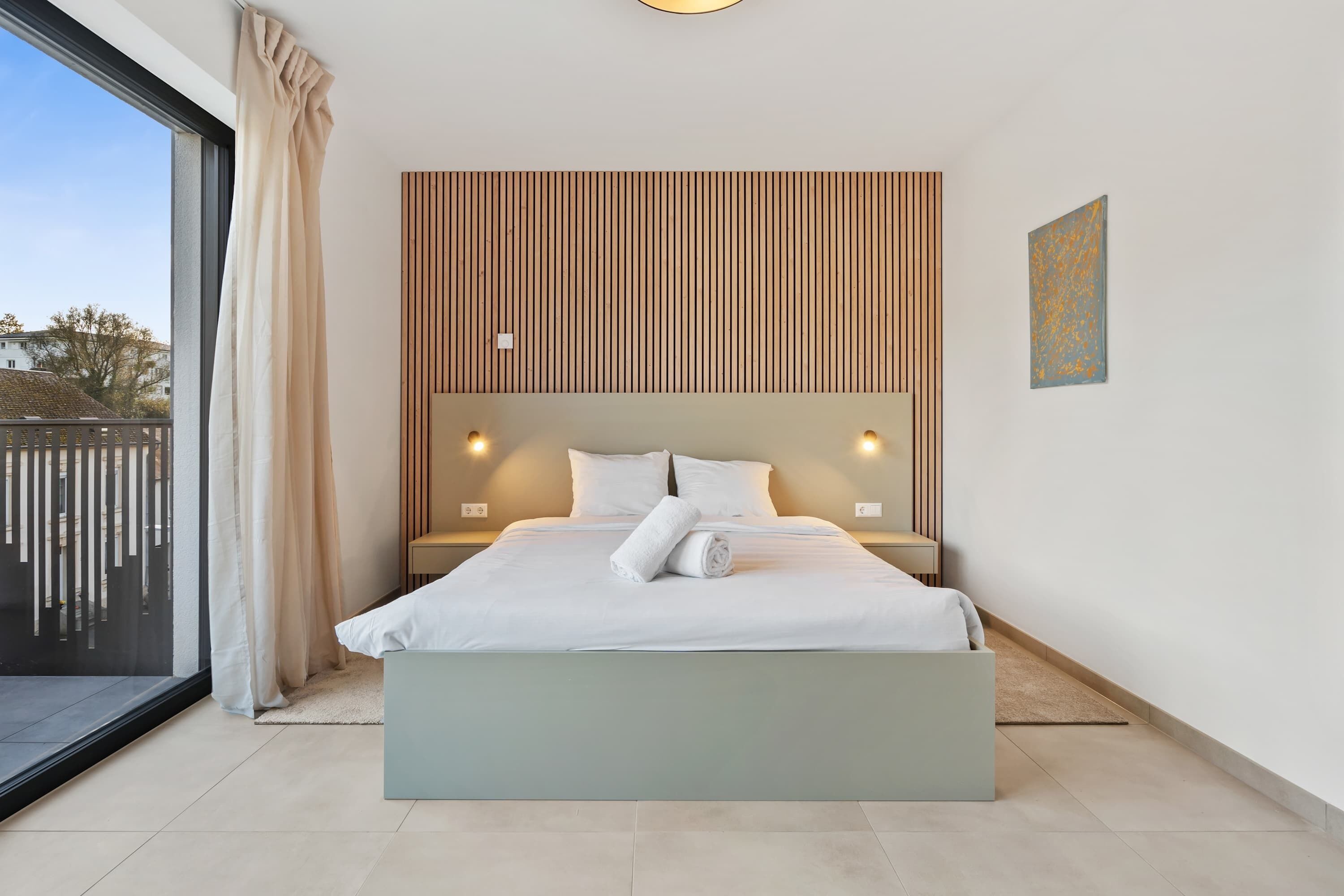 A modern bedroom with a large bed, a view through a sliding glass door, and a wooden slat wall design.