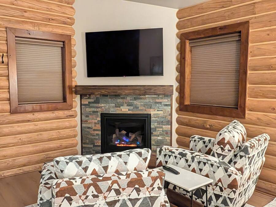 A log cabin interior with a fireplace, two patterned armchairs, a small table, and a mounted TV.