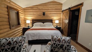 Een slaapkamer in de stijl van een blokhut, met een bed, twee bedlampjes, een hoofdbord met een hondenhoofd en een grote houten deur.