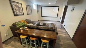 A modern living room with a sectional sofa, a wooden dining table with stools, and a flat-screen TV mounted on the wall. - Pool! Arcade! Theatre Room! Firepit! 4 BR (Lincoln)
