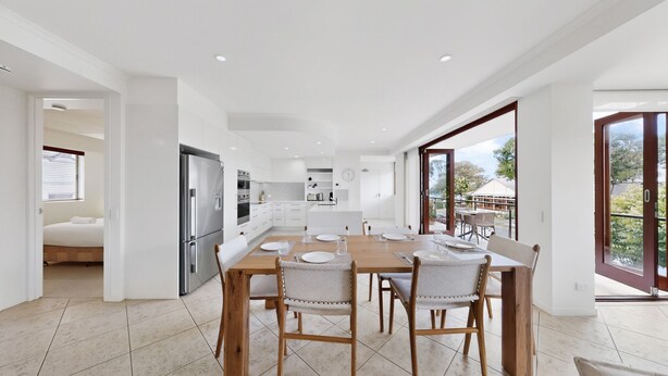 A modern kitchen with white cabinetry, a dining area with a wooden table and chairs, and large sliding glass doors leading to a balcony. - Beautiful and spacious 3 bedroom apartment (Noosaville)