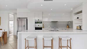 A modern kitchen with white cabinetry, a central island with bar stools, a built-in oven, and a tiled backsplash. - Beautiful and spacious 3 bedroom apartment (Noosaville)