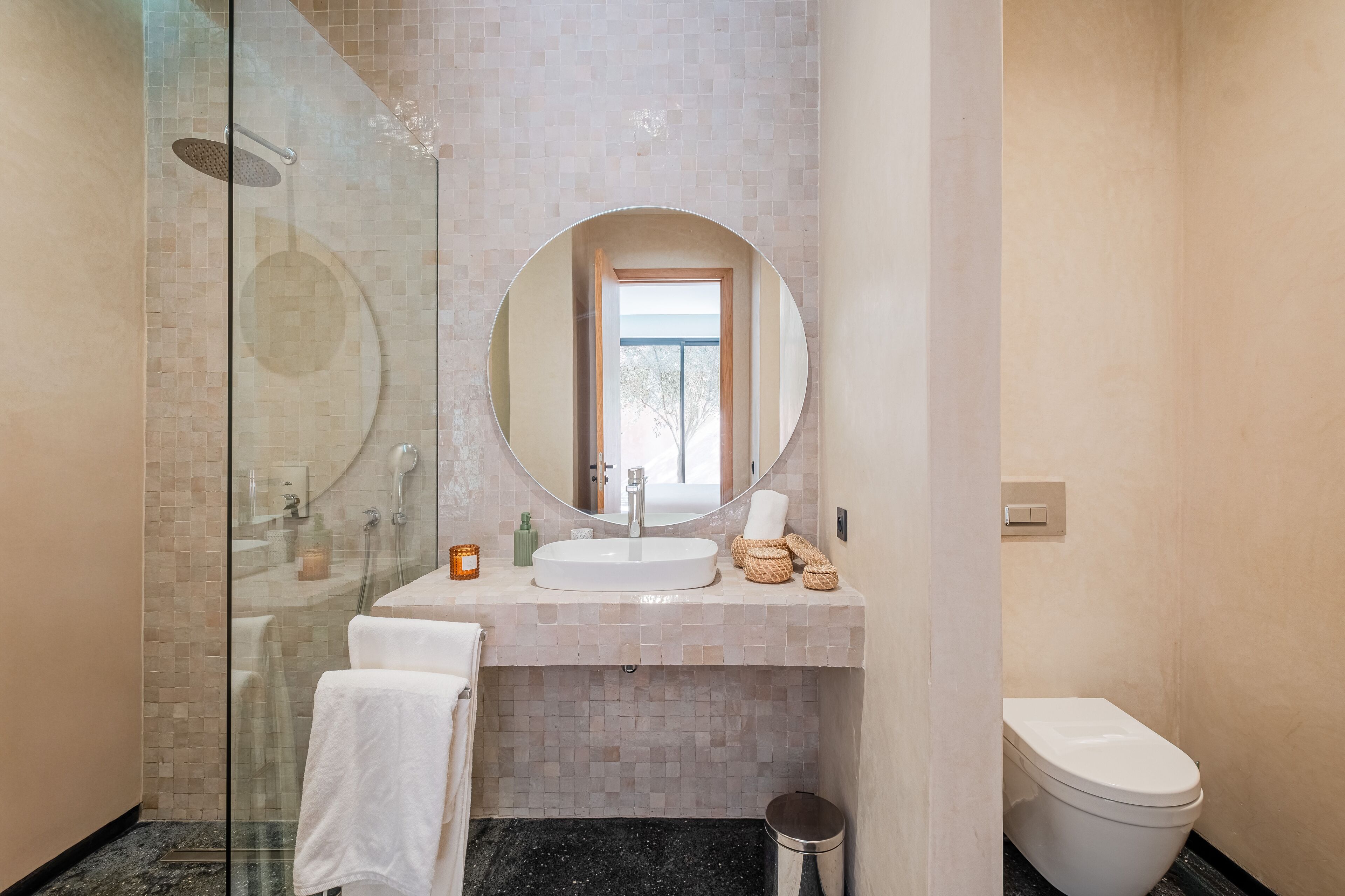 A modern bathroom with a glass-enclosed shower, a round mirror, a sink, and a toilet.