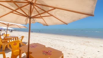 Area salotto in spiaggia con sedie e tavoli gialli sotto ombrelloni beige, con l'oceano e la spiaggia sullo sfondo.