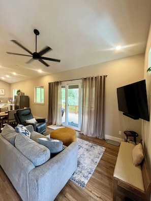 Un salon moderne avec un canapé gris, un fauteuil en cuir noir, un ventilateur de plafond et un téléviseur fixé au mur.