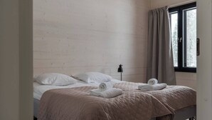 Et værelse med to senge, hver med et quiltet sengetæppe og puder. Der er et vindue med gardiner, en lampe på et stativ og udsigt til træer udenfor.